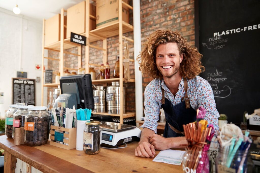 A shop worker with curly hair and an apron stands behind a counter with jars, a scale, and supplies in a plastic-free store.