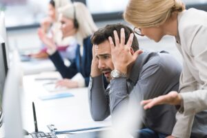 A man at a desk looks stressed, holding his head, while a woman stands beside him talking; other people work at computers in the background.